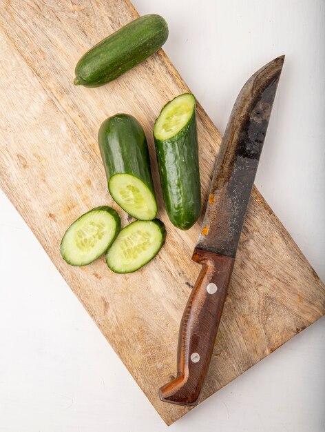 Side view of whole and cut cucumbers with knife on cutting board on white surface