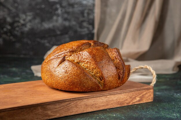 Side view of whole fresh black bread on brown wooden cutting board on dark surface