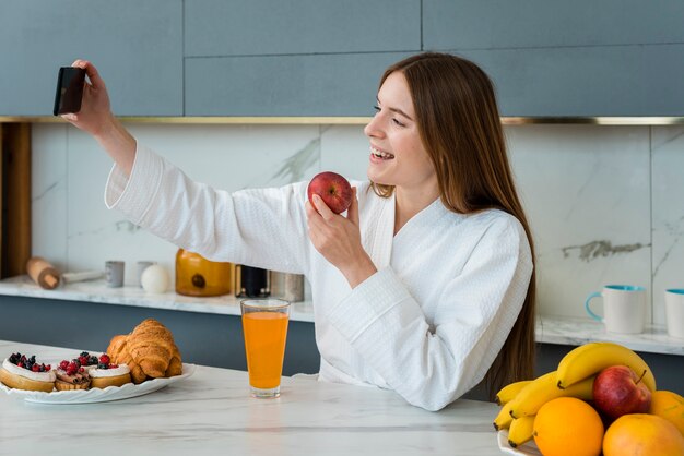 Side view of woman in bathrobe taking a selfie and holding apple