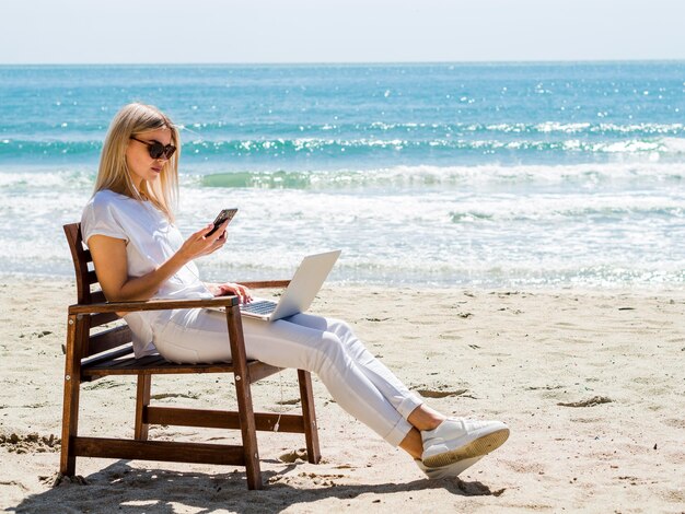 Foto gratuita vista laterale della donna nella sedia di spiaggia con il computer portatile e lo smartphone