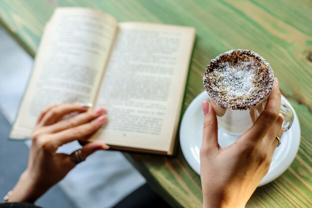 Вид сбоку женщина пьет латте с кокосом и читает книгу за столом
