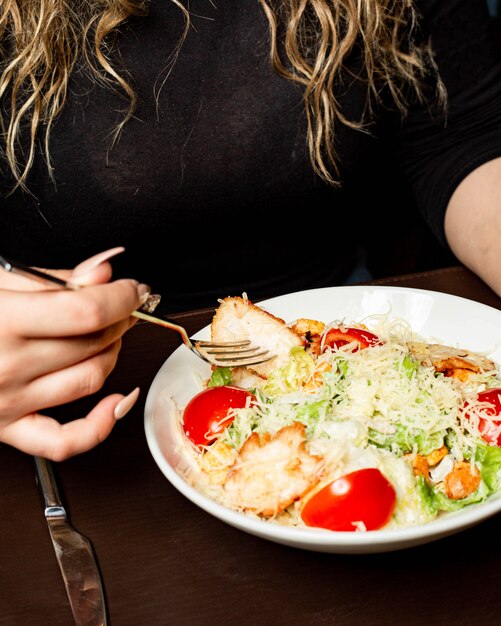 Side view of a woman eating caesar salad with chicken and parmesan cheese in bowl at the table
