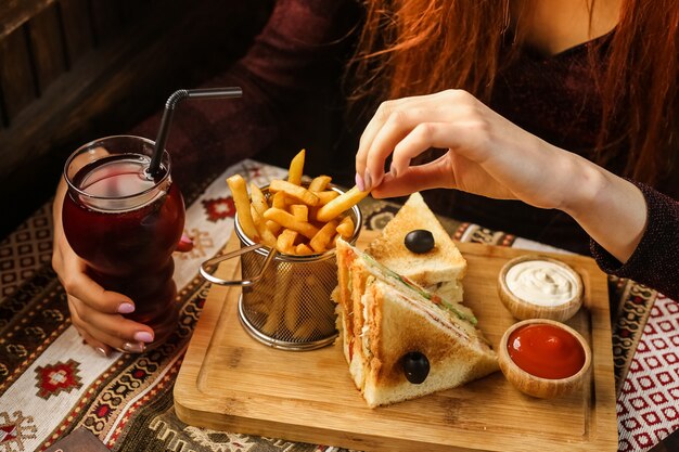 Side view woman eating french fries with club sandwich ketchup and mayonnaise on stand with soft drink