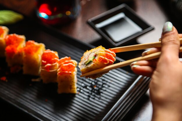 Side view woman eating fried sushi in sauce with chopsticks and soy sauce on a stand