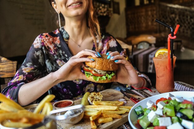Вид сбоку женщина ест мясной бургер с картофелем фри кетчупом и майонезом на деревянной подставке