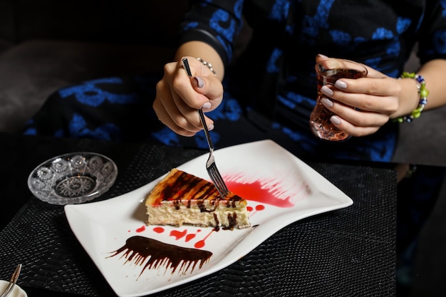 Free photo side view a woman eats cheesecake with chocolate syrup and drinks tea in an armudu glass