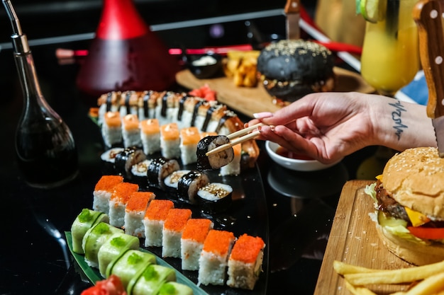 Side view woman eats mix sushi rolls with soy sauce and burgers on the table