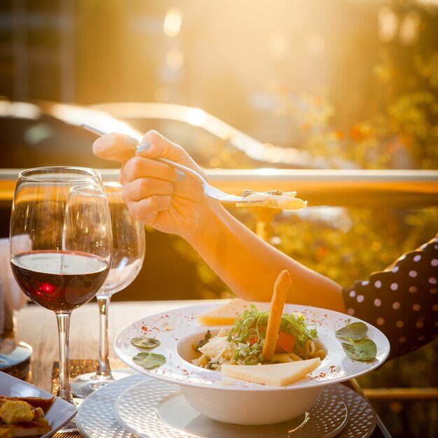 Side view woman eats salad with a glass of wine on table with trees and sunlight leak on background