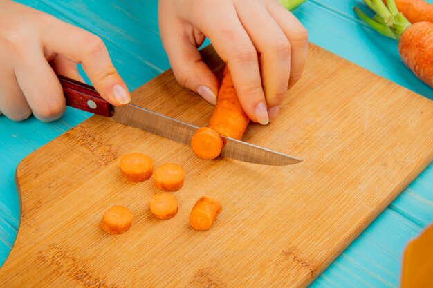 Вид сбоку женщина руки резки морковь на разделочную доску с ножом на синем фоне