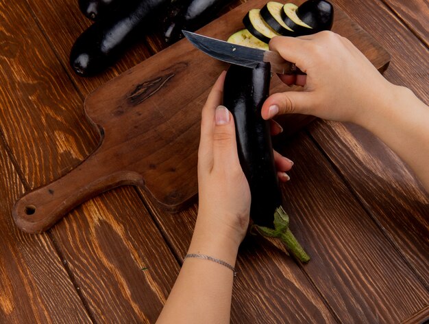 Side view of woman hands cutting eggplant with knife on cutting board with whole ones on wooden background