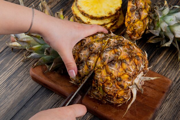 Side view of woman hands cutting pineapple with knife on cutting board with sliced pineapple on wooden background