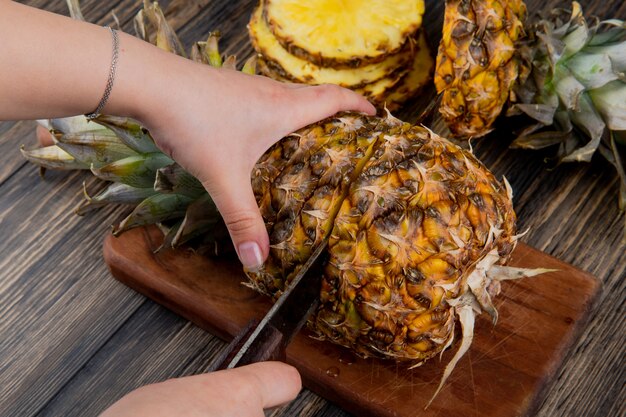 木製の背景にスライスしたパイナップルとまな板の上のナイフでパイナップルを切る女性の手の側面図