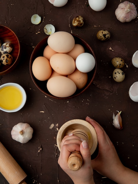 Вид сбоку женских рук отжимать чеснок в чесночной дробилке с яйцом и маслом вокруг на бордовом фоне