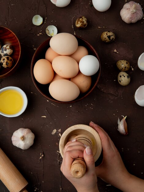 Side view of woman hands pressing garlic in garlic-crusher with eggs and butter around on maroon background