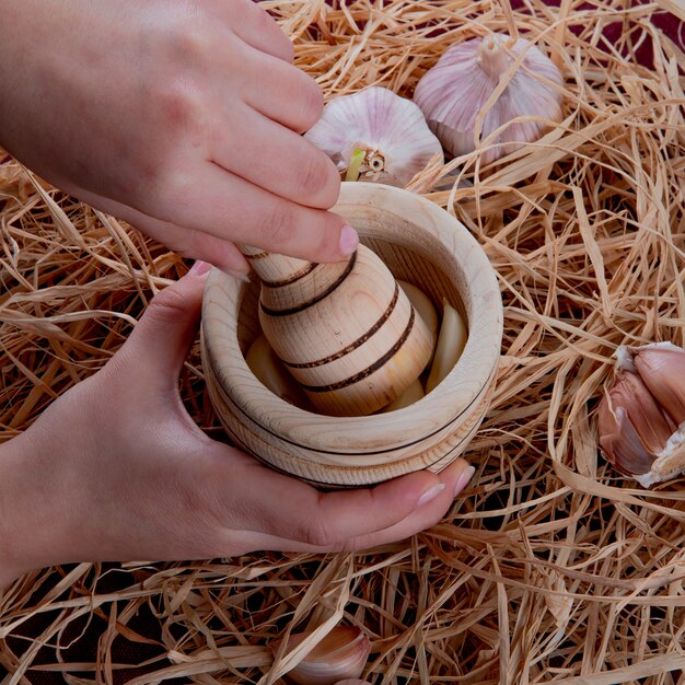 Side view of woman hands pressing garlic in garlic crusher with garlic bulbs around on straw background