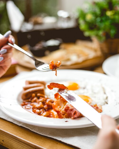 Side view a woman has breakfast fried eggs with sausages beans and bacon on a plate