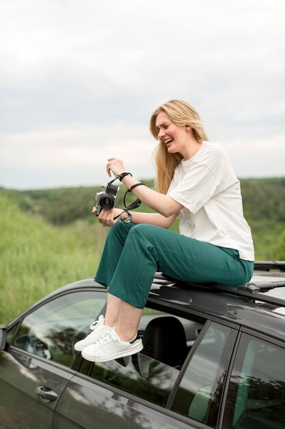 Side view of woman holding camera while posing on top of car