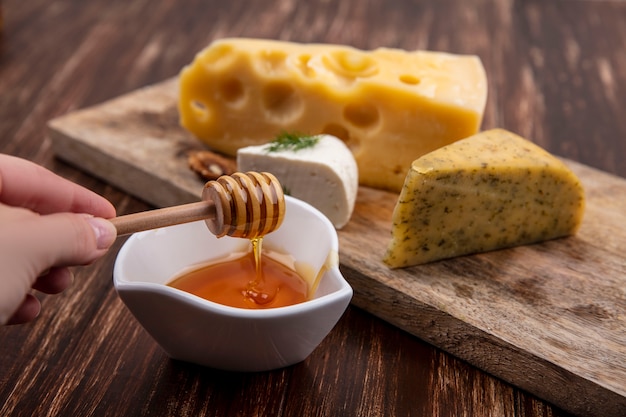 Free photo side view  a woman holds a wooden bag with honey with a saucer with varieties of cheeses on a stand  on a wooden background