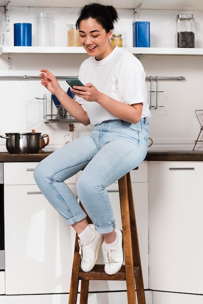 Вид сбоку женщины дома на кухне с помощью смартфона