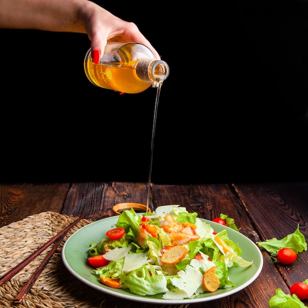 Side view woman pouring oil on delicious salad in plate on wooden and black background.