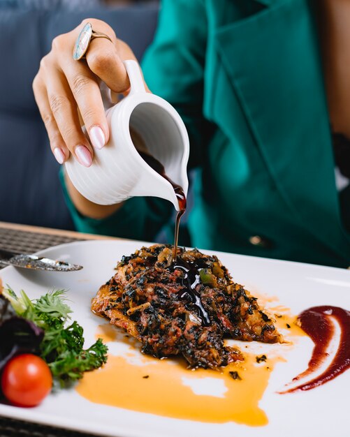 Side view of a woman pouring pomegranate narsharab sauce on fried fish fillet with vegetables on a plate