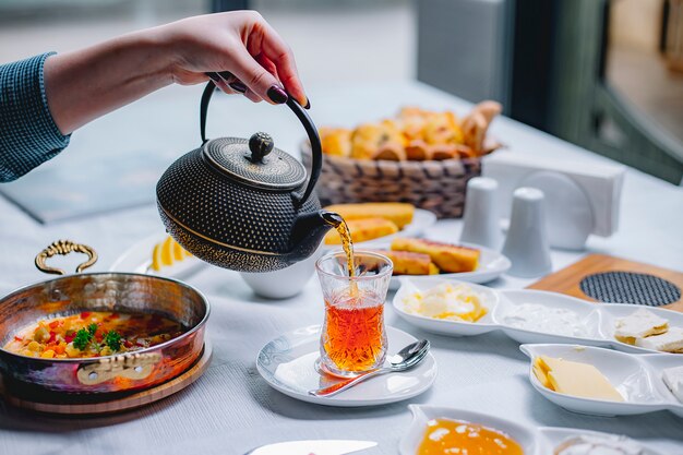 Side view a woman pours in a glass of armudu tea with a served breakfast on the table