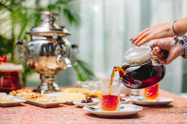 Side view a woman pours tea from a teapot into a glass of armuda on a saucer