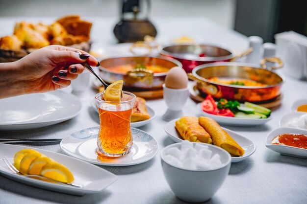 Side view woman put in cup of tea lemon slice pancakes with boiled eggs tomatoes cucumbers and honey on the table serve breakfast