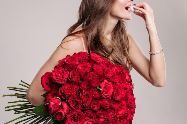 Side view of woman's body in move eating white coconut candy Brunette girl with long hair and elegant bracelet on hand holding bouquet of red flowers Concept of holiday woman's presents and jewelry