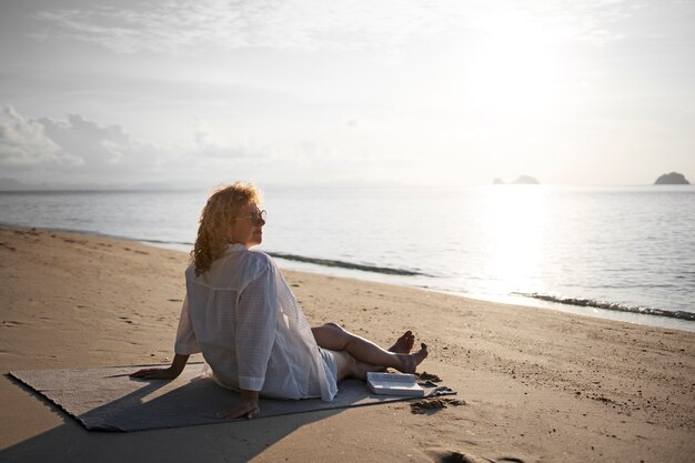 Вид сбоку женщина сидит на пляже с книгой