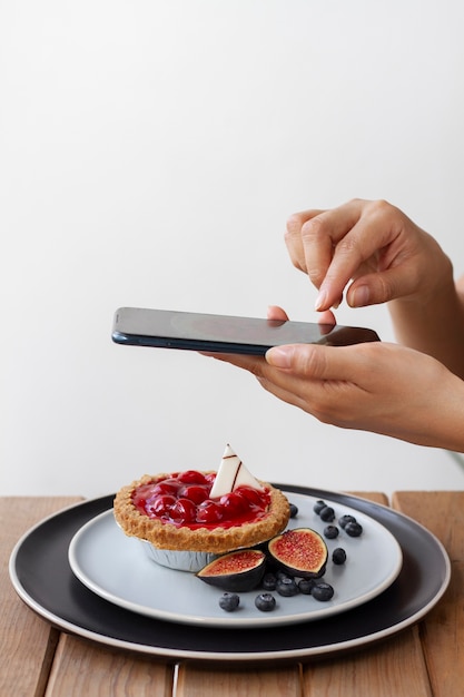 Side view of woman taking photo of fruit tart with smartphone