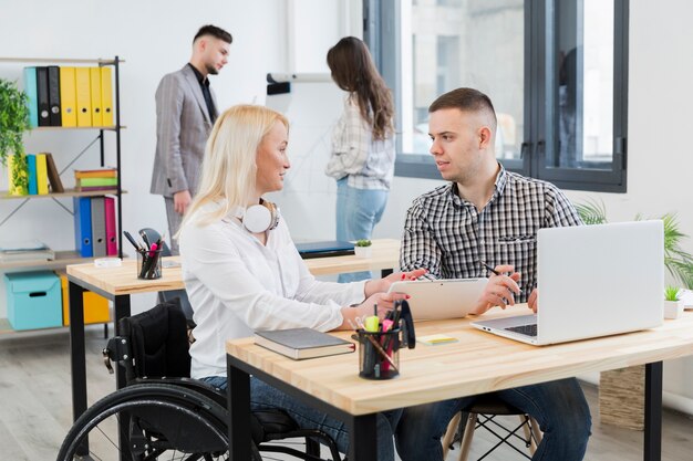 Side view of woman in wheelchair discussing with coworker at her desk