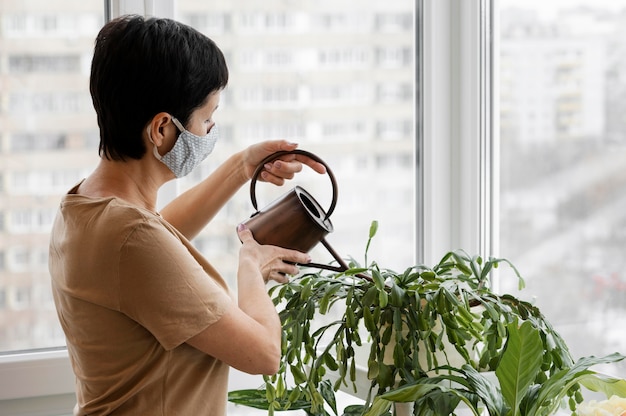 Вид сбоку женщины с маской для лица, поливающей растения в помещении