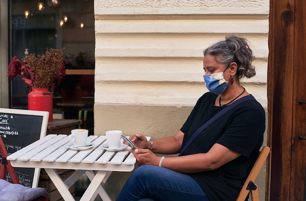 カフェの屋外テーブルで彼女の携帯電話を使用しながらフェイスマスクを持つ女性の側面図