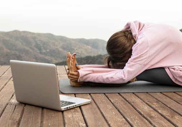 Вид сбоку женщины с ноутбуком, занимающейся йогой на открытом воздухе