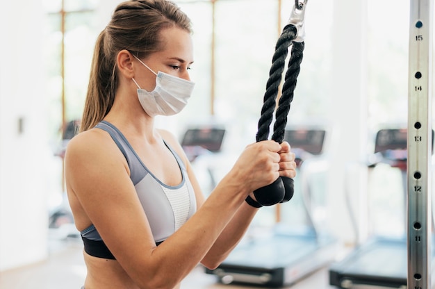 Вид сбоку женщины с медицинской маской, тренирующейся в тренажерном зале во время пандемии