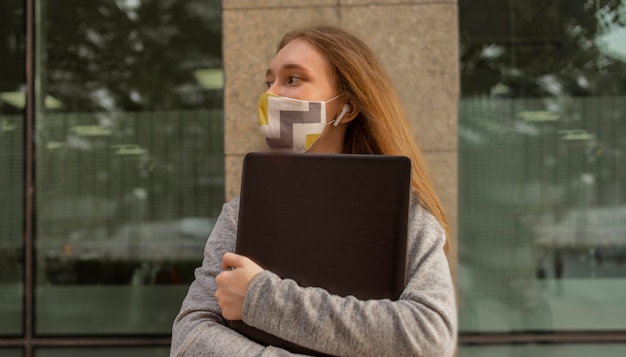 Side view woman with medical mask holding her laptop outside