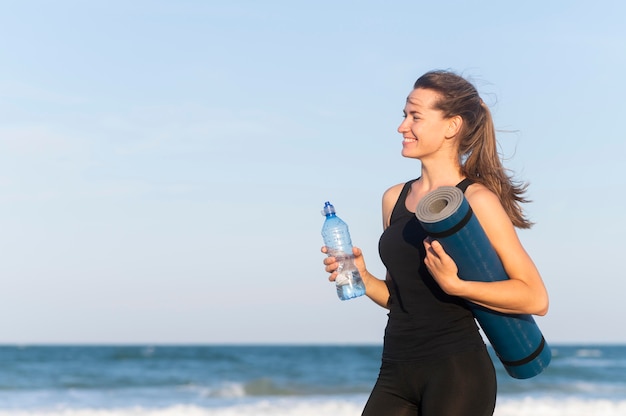 水のボトルとビーチでヨガマットと女性の側面図