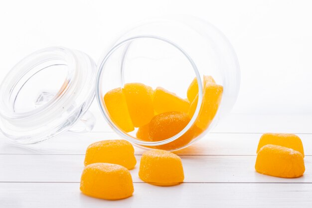 Side view of yellow marmalade candies scattered from a glass jar on white