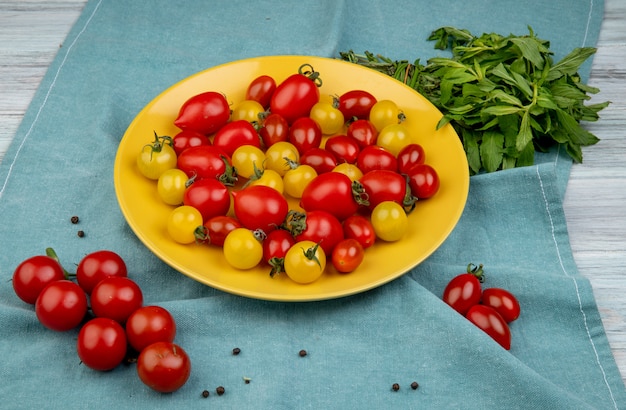 Side view of yellow and red tomatoes in plate and green mint leaves on blue cloth surface