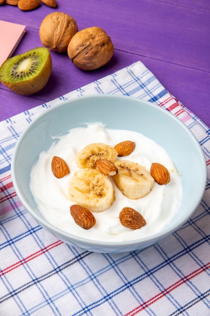 Free photo side view of yogurt with sliced bananas and almond in a white bowl, kiwi fruit and walnuts on purple wood