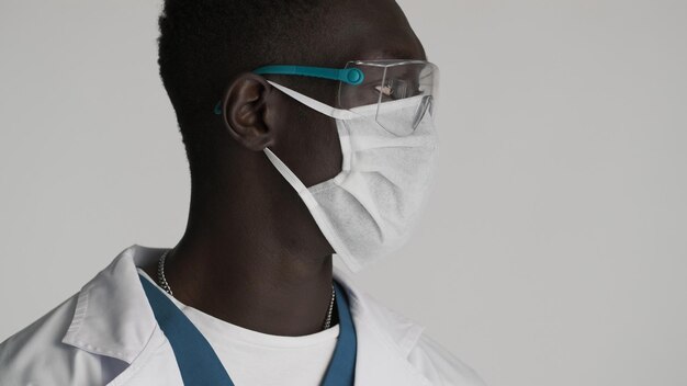 Side view of young African American male doctor in protective eyeglasses and medical mask looking confident over white background