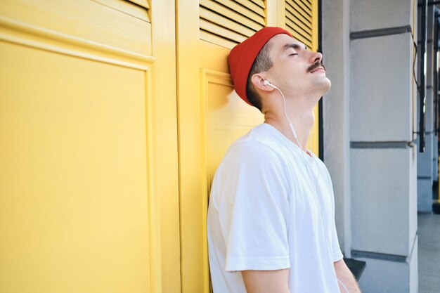 Side view of young attractive mustache man in T-shirt and red hat dreamily listening music in earphones with yellow background on city street