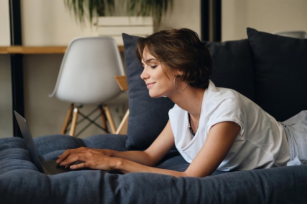 Vista laterale della giovane donna attraente sdraiata sul divano che lavora felicemente su un nuovo progetto con il laptop in uno spazio di coworking moderno