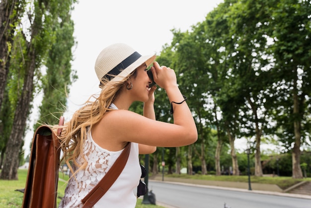 Free photo side view of a young blonde woman wearing hat taking photograph on camera