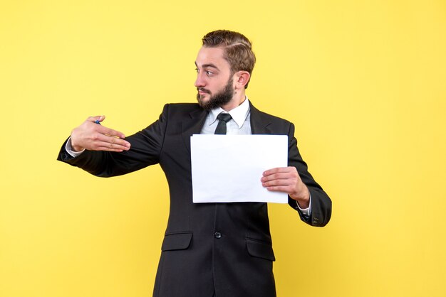 Side view of young businessman looks aside and pointing with a right hand to a blank paper on the yellow wall