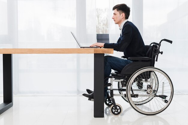 Side view of a young businessman sitting on wheelchair using laptop in new office