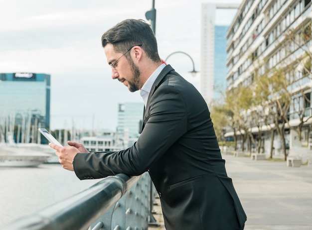 Side view of a young businessman standing near the railing using smartphone