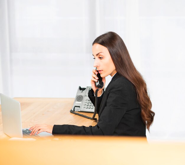 Взгляд со стороны молодой коммерсантки используя компьтер-книжку пока говорящ на телефоне в офисе