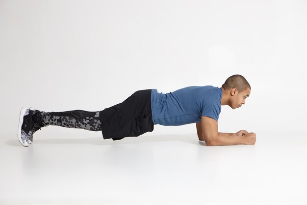 Side view of young dark skinned sportsman doing elbow plank in studio wearing stylish sneakers and training clothes, looking ahead of him with determined concentrated expression. Endurance and stamina